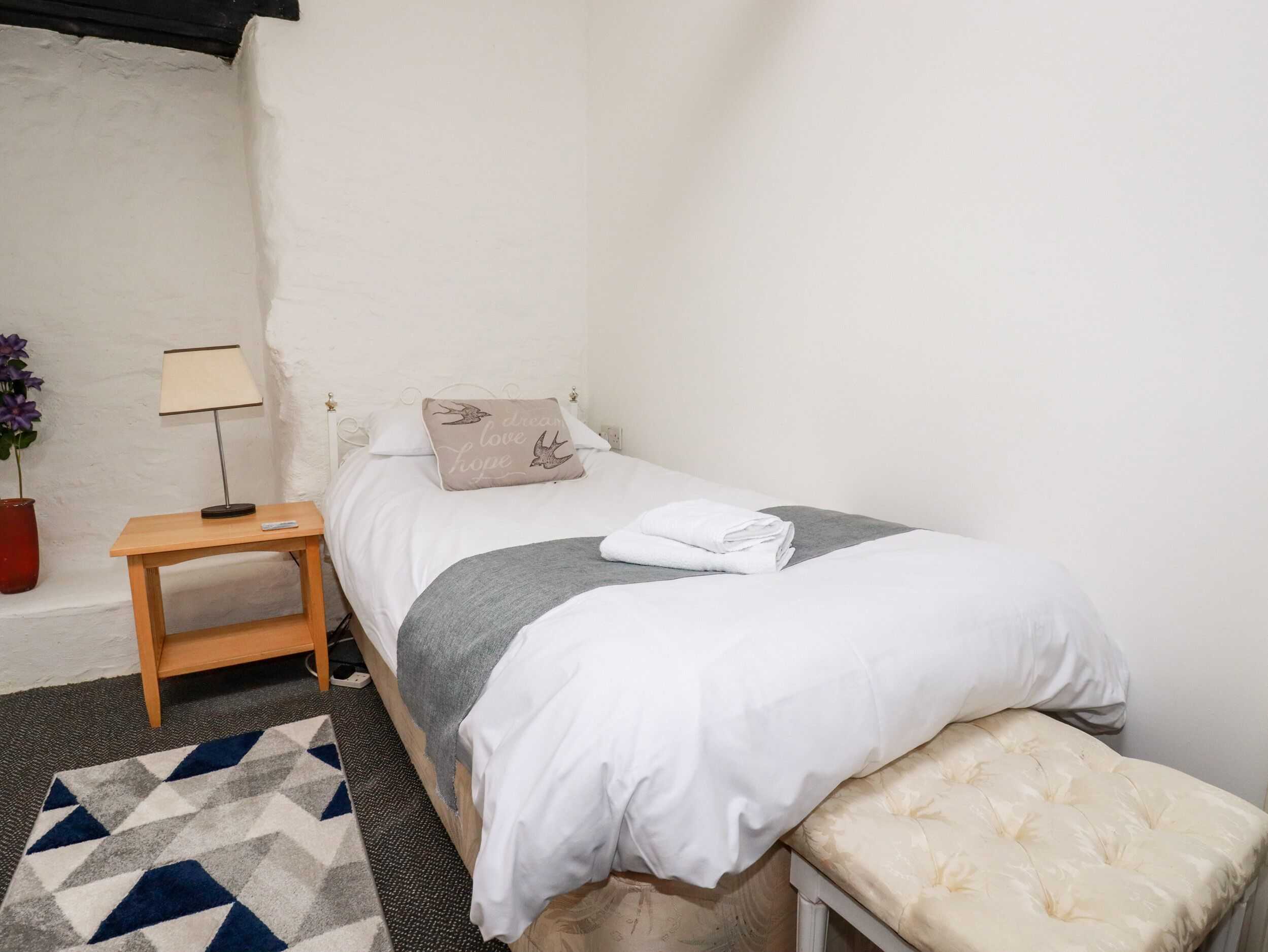 Une petite chambre à coucher, bien rangée, avec un lit simple, une table de chevet, une lampe et un petit banc.