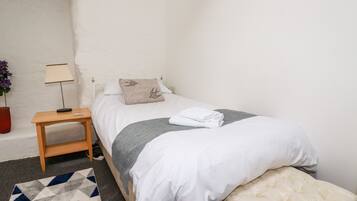 Une petite chambre à coucher, bien rangée, avec un lit simple, une table de chevet, une lampe et un petit banc.