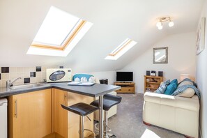 A compact kitchen with a central island, a dining area with bar stools, a sofa, and a TV. - The Litte Hideaway (Newquay)