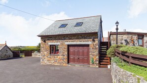 A stone building with a brown door and windows, a paved driveway, and a stone wall with greenery. - The Litte Hideaway (Newquay)