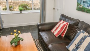 En stue med en lædersofa, et træsofabord med en potteplante, og udsigt til en kystby gennem store vinduer.