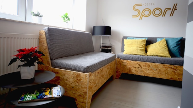Una habitación moderna con un sofá de madera, una silla más pequeña, una mesa con una revista, una planta en maceta y una lámpara. La habitación tiene un ventanal grande y un letrero en la pared que dice 'Hostel Sport'.