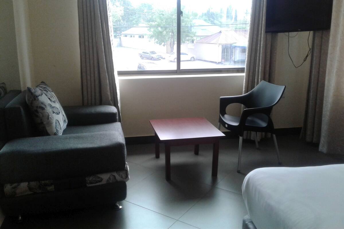 Una habitación de hotel con cama, sofá, sillón, una mesita y televisor montado en la pared.