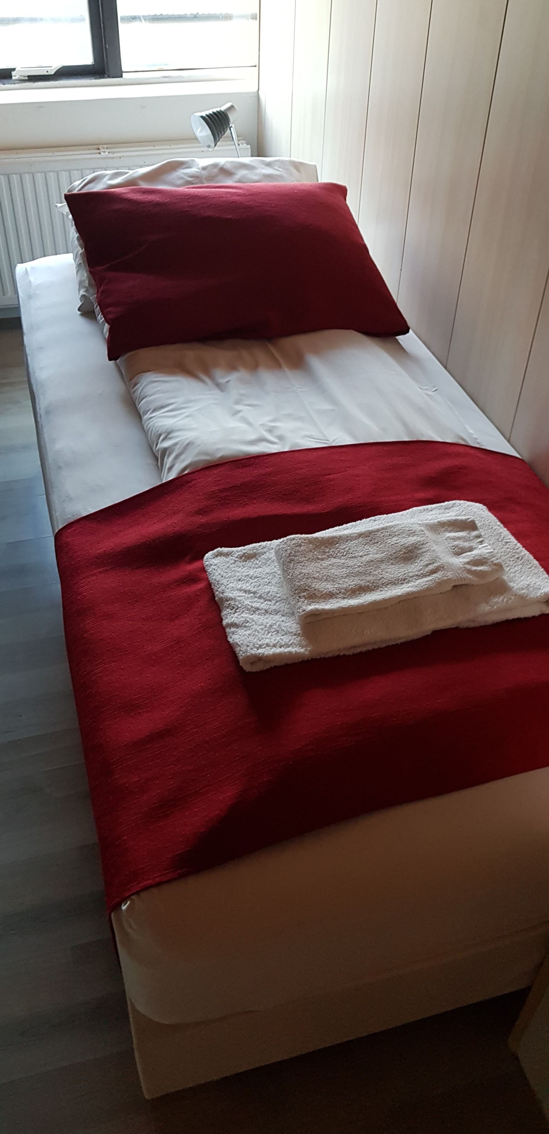 A single bed with red and white bedding, a white towel, and a lamp on a bedside table.