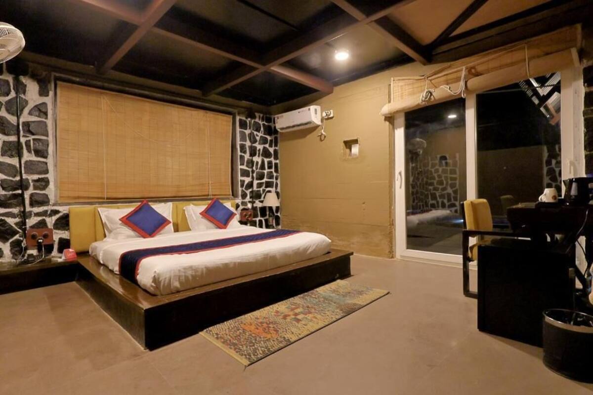 A bedroom with a bed, a nightstand, a lamp, a fan, a window with blinds, and a view of the outside.