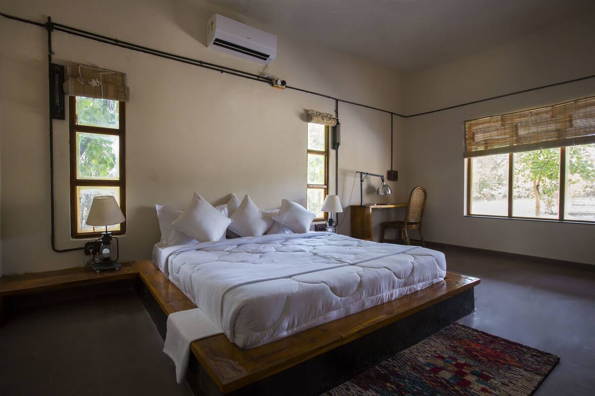 A bedroom with a large bed, a bedside table with a lamp, a chair, and a window with a view of greenery.