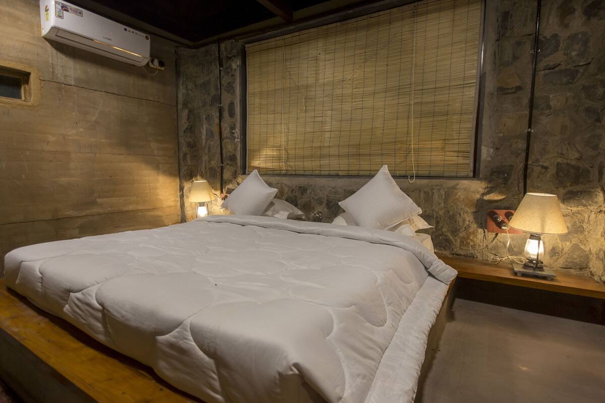 A bedroom with a large bed, two bedside tables with lamps, a wall-mounted air conditioner, and a window with blinds.