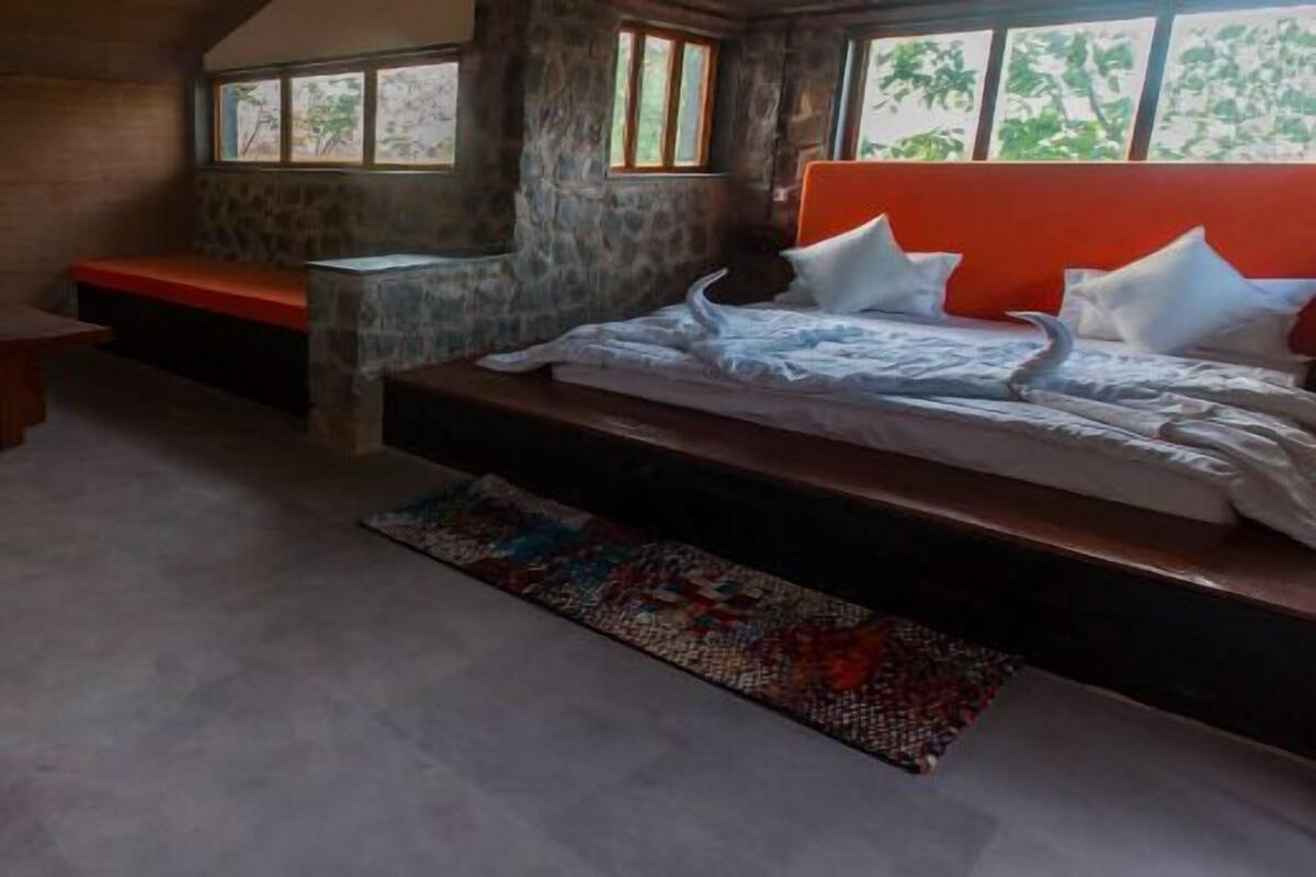 A bedroom with a stone wall, a wooden bench, a bed with white bedding, and a colorful rug.