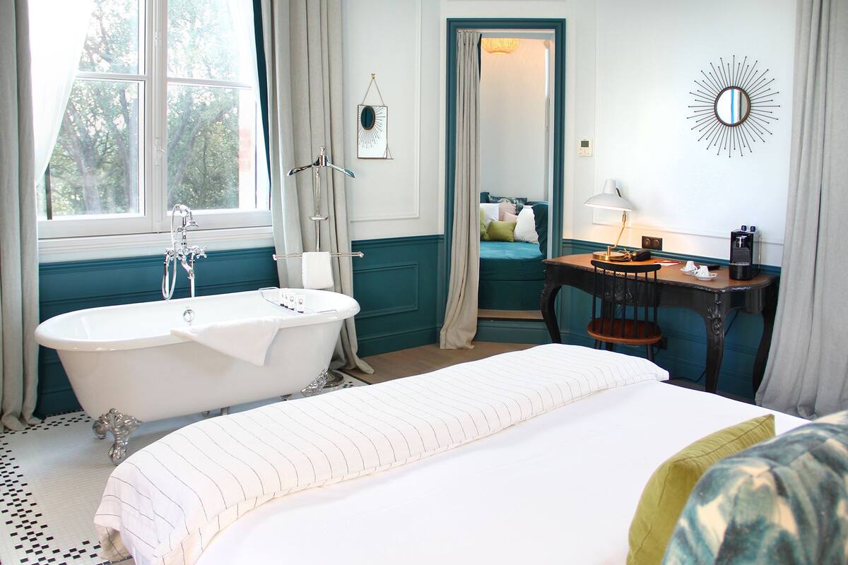 A room with a large bed, a bathtub, a desk with a lamp, and a mirror on the wall.