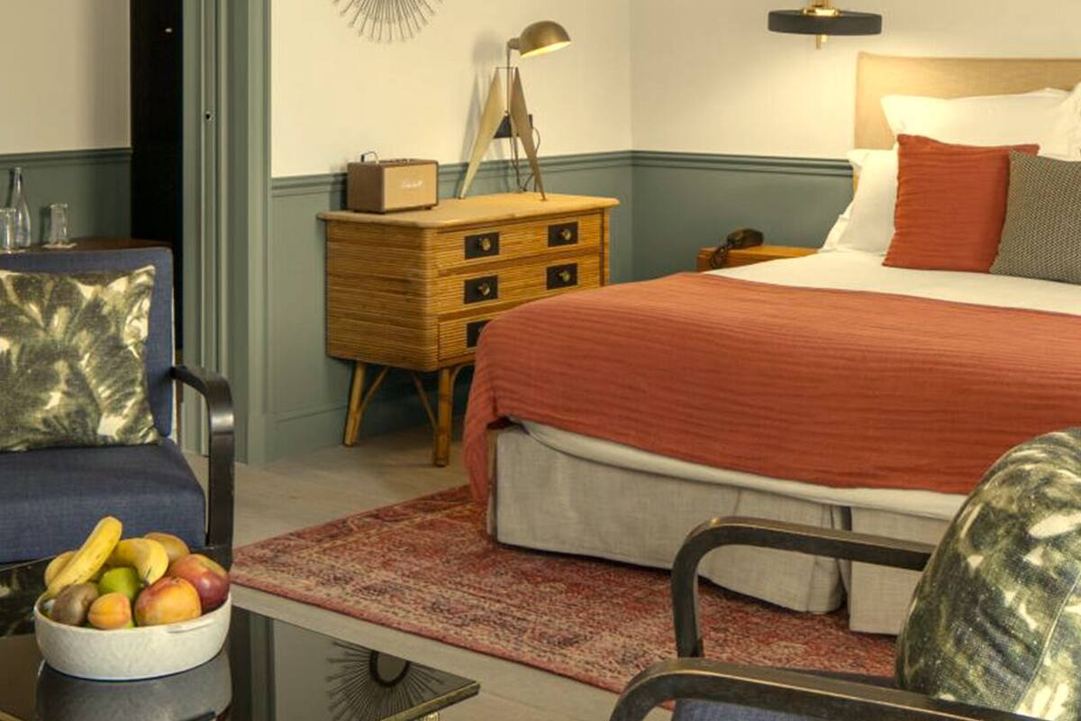 A hotel room with a bed, a desk with a bowl of fruit, a chair, a dresser, and a wall-mounted lamp.