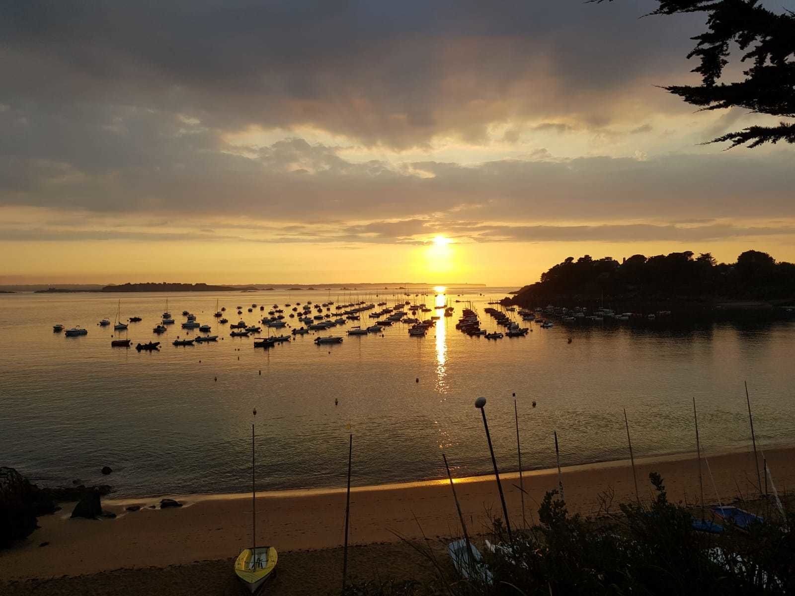 Una puesta de sol sobre un mar tranquilo con barcos anclados en fila, una playa arenosa en primer plano y una costa lejana con árboles.