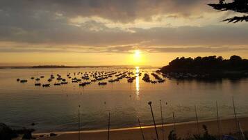 Una puesta de sol sobre un mar tranquilo con barcos anclados en fila, una playa arenosa en primer plano y una costa lejana con árboles.