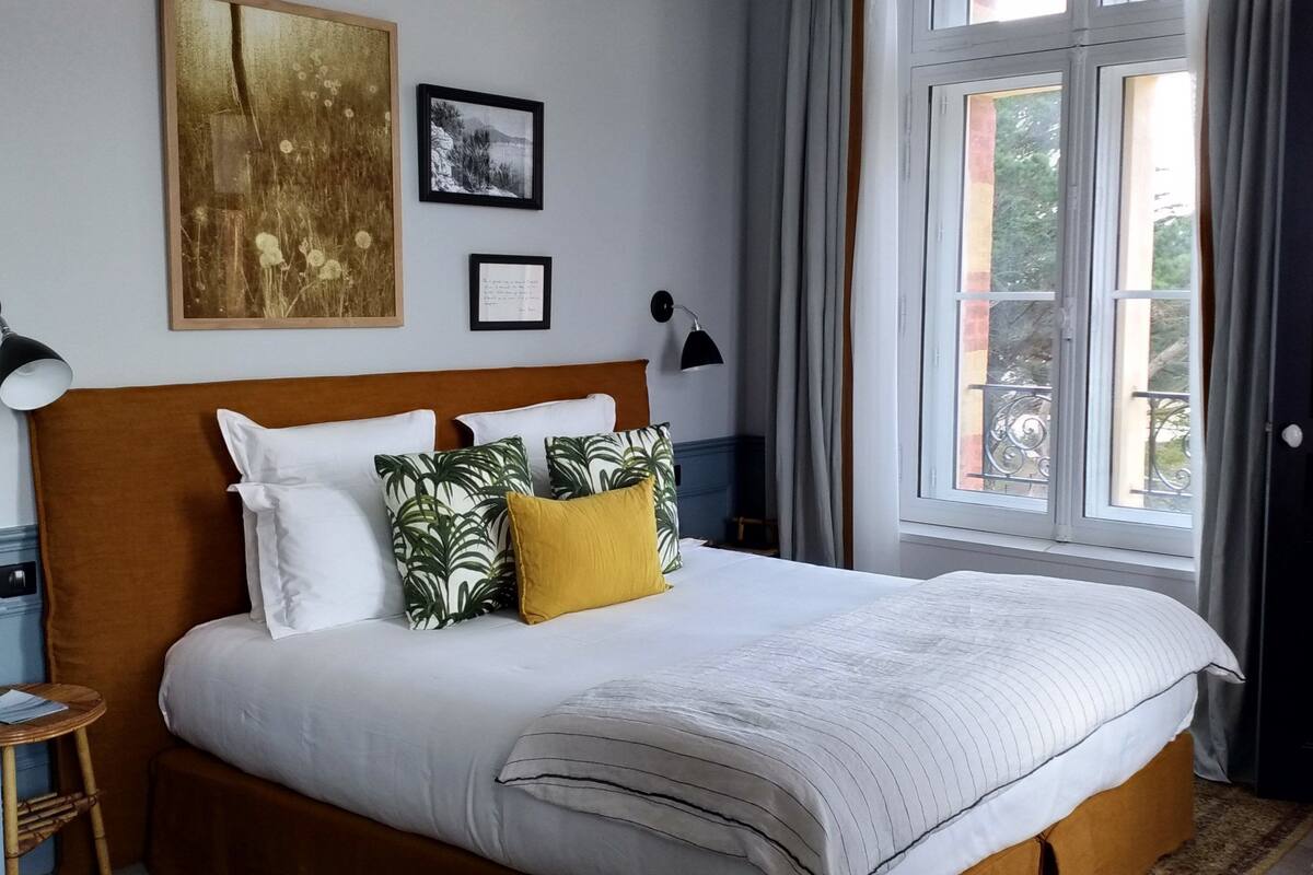 A neatly arranged bedroom with a large bed, a bedside table, a lamp, and a view of the outdoors through a window.