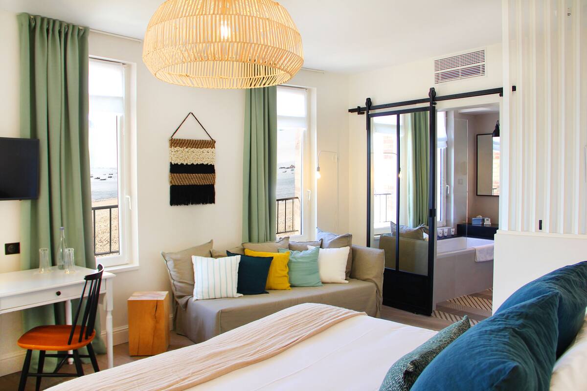 A hotel room with a bed, a desk, a chair, a sofa, and a bathroom visible through a sliding door.