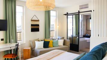 A hotel room with a bed, a desk, a chair, a sofa, and a bathroom visible through a sliding door.