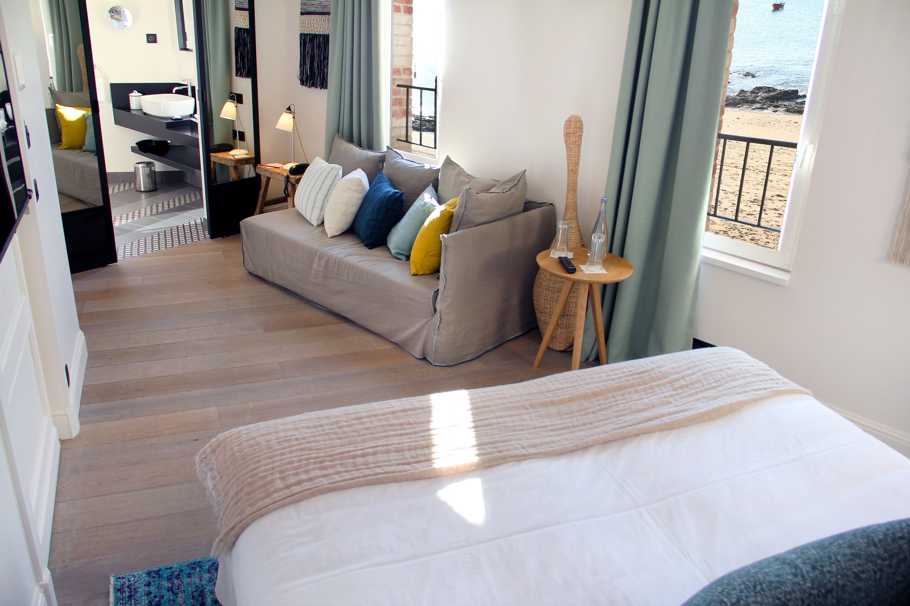 A hotel room with a bed, a sofa, a small table with a lamp, a window with a view of the beach, and a bathroom with a sink and mirror.
