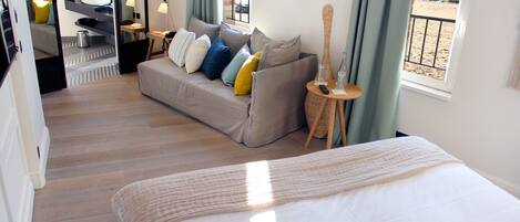 A hotel room with a bed, a sofa, a small table with a lamp, a window with a view of the beach, and a bathroom with a sink and mirror.