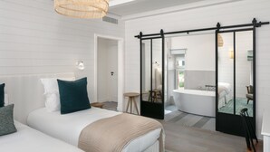 A modern hotel room with a large bed, a sitting area with a chair and a small table, a bathroom with a bathtub, and a sliding door leading to a balcony. - Le Nessay (Saint-Briac-sur-Mer)