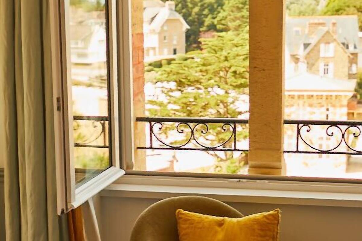 A room with a bed, a window with curtains, a chair with a yellow pillow, and a view of buildings and greenery outside.