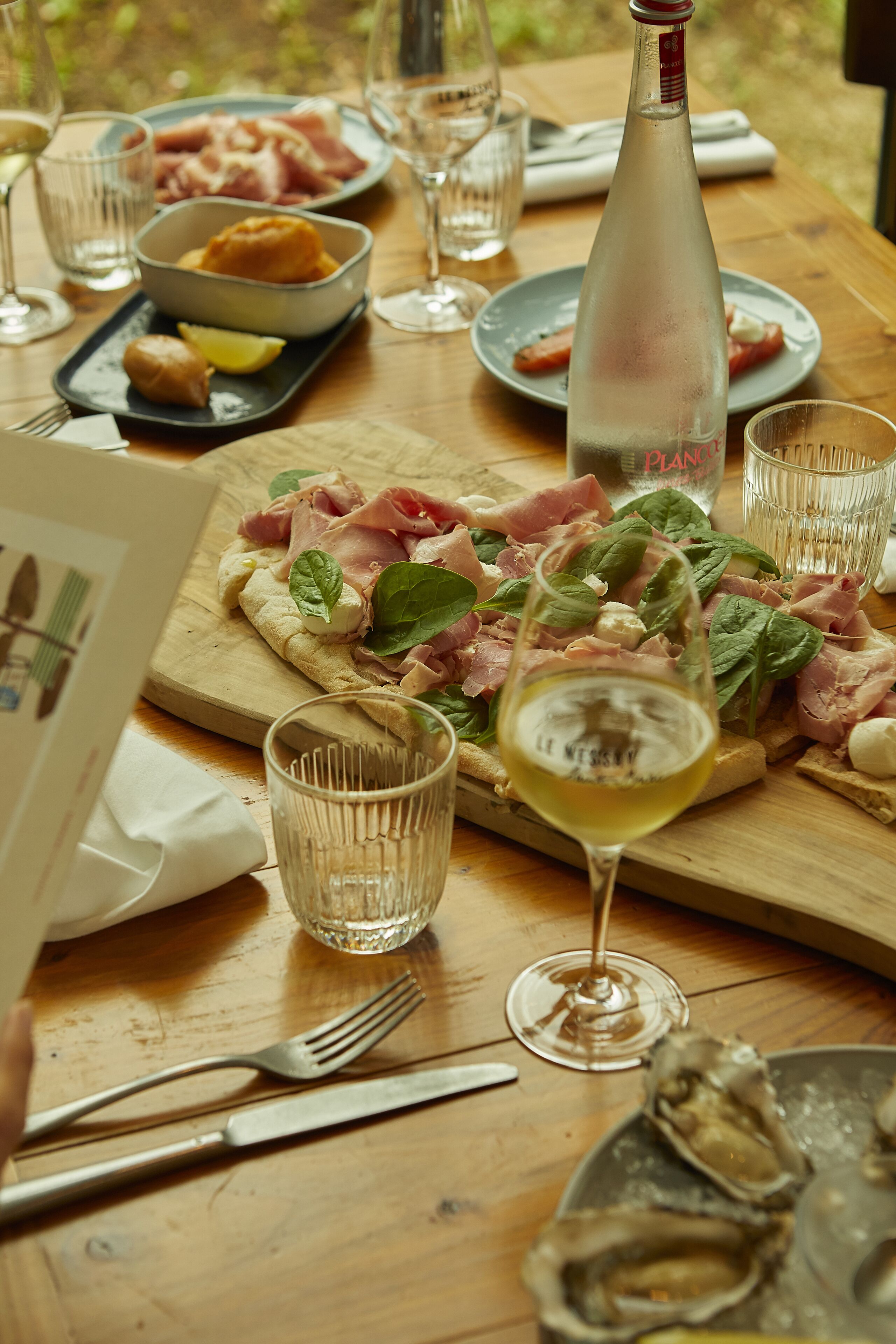 A wooden table with a meal, including a pizza with ham and cheese, a bottle of water, and a glass of white wine.