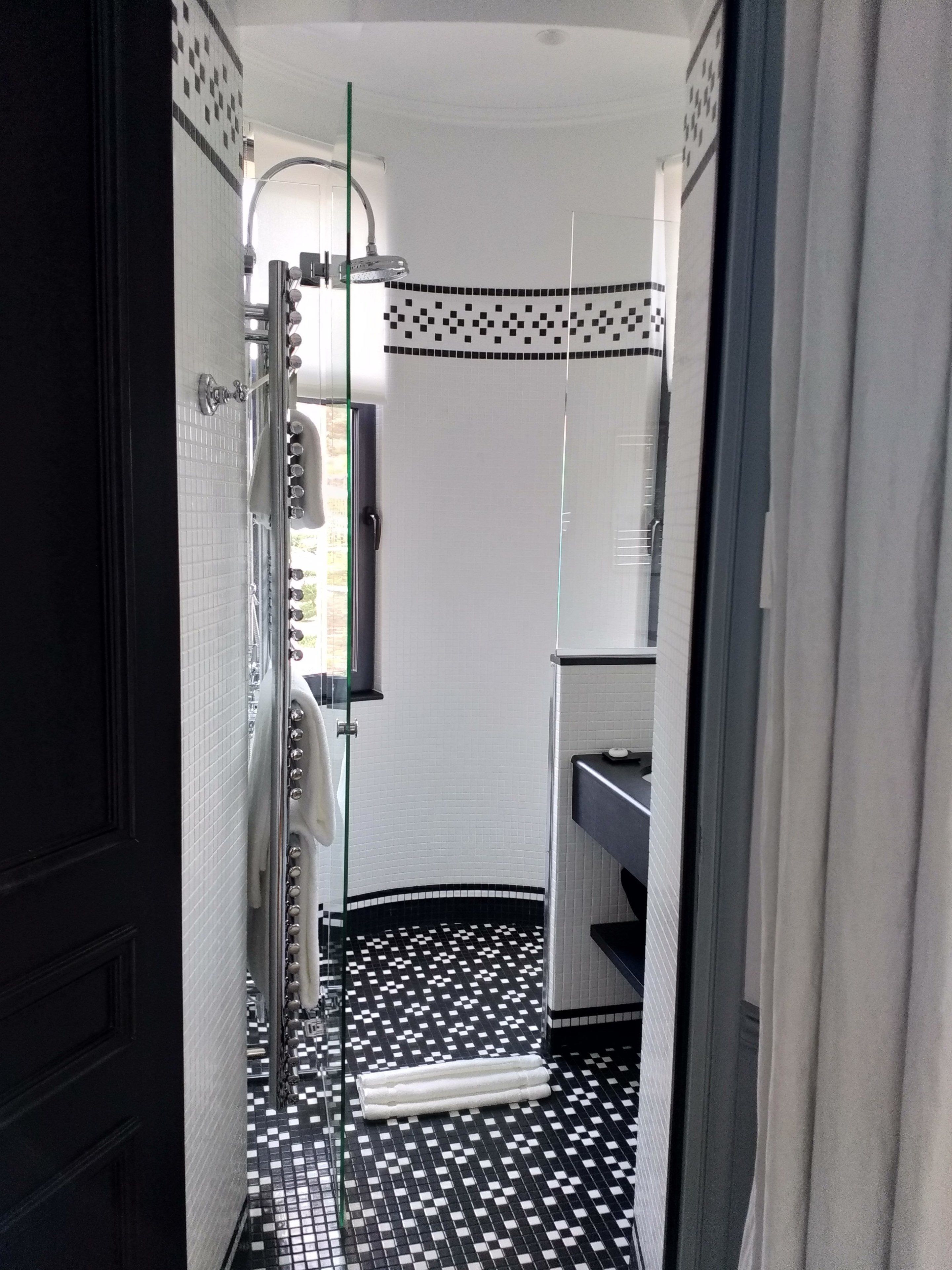Un couloir étroit doté d’un sol à carreaux noirs et blancs, d’une cabine de douche en verre et d’un mur recouvert de carreaux blancs agrémenté d’une bordure décorative.