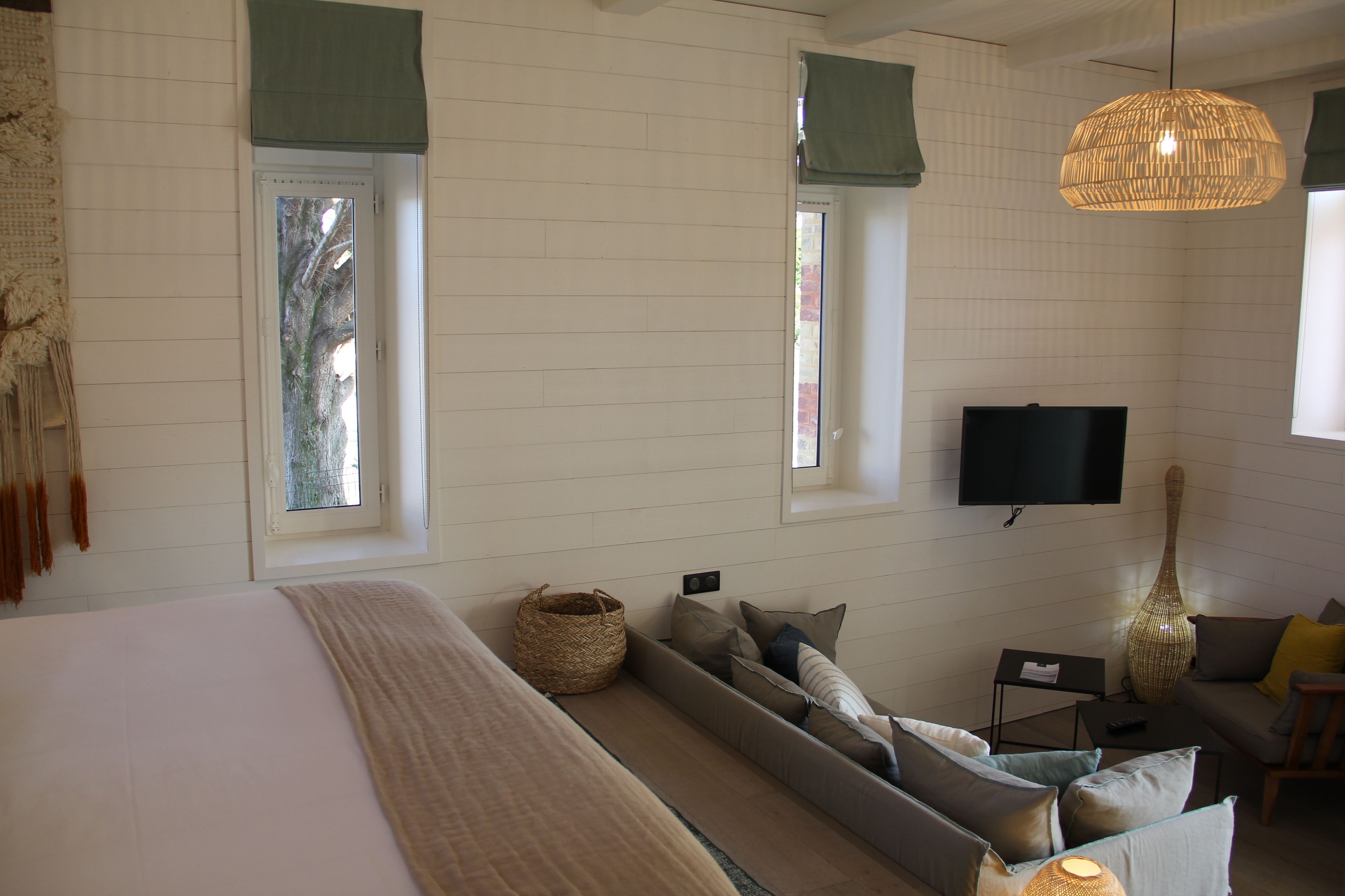 A modern bedroom with a large bed, a sofa bed, a flat-screen TV, and a large window with a view of trees.