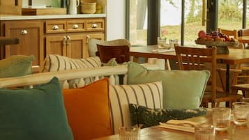 A dining area with wooden tables and chairs, a bench with cushions, and a shelf with glassware.