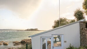 A coastal building with a white exterior, a door leading to an interior with a table and chairs, and a view of the sea and boats in the background. - Le Nessay (Saint-Briac-sur-Mer)