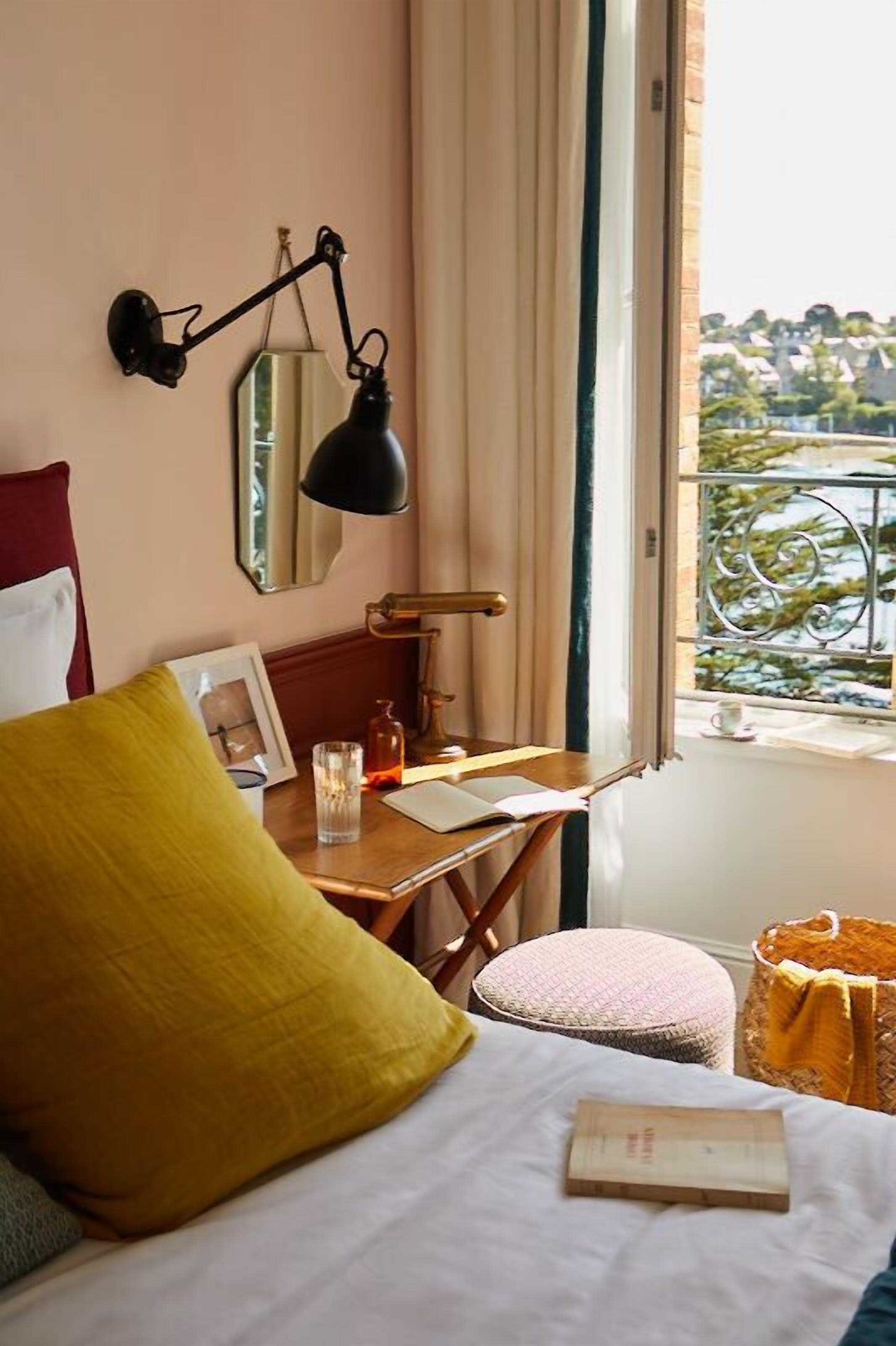 A hotel room with a bed, a bedside table with a lamp, a window with a view of greenery, and a small round ottoman.