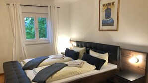 A neatly made bed with pillows and a blanket, a bedside table with a lamp, and a window with curtains. - Alpenlodge (Garmisch-Partenkirchen)