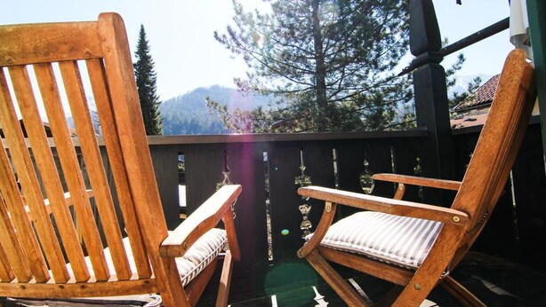 Two wooden chairs with cushions on a balcony with a view of trees and mountains. - Alpenlodge (Garmisch-Partenkirchen)