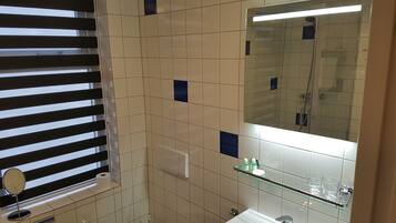 Une salle de bain avec un grand miroir, une étagère en verre où sont rangés les articles de toilette et une fenêtre avec des stores.