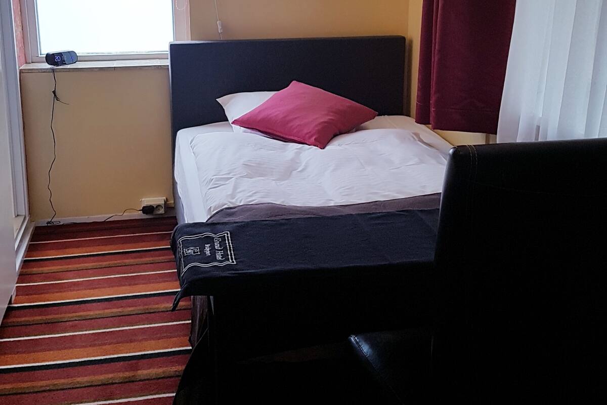 A hotel room with a bed, a chair, a window with curtains, a wall-mounted picture, and a chandelier.