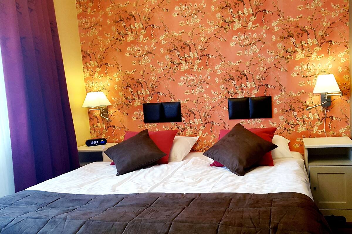 A hotel room with a floral wallpaper, a bed with multiple pillows, bedside lamps, and a nightstand with a clock.