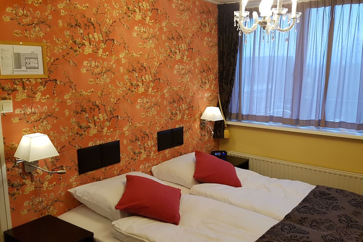A hotel room with a floral wallpaper, two beds with red and white bedding, a chandelier, a window with sheer curtains, and a wall-mounted lamp.