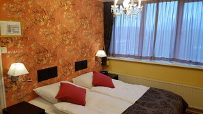 Una habitación de hotel con papel tapiz floral, dos camas con ropa de cama roja y blanca, una araña, una ventana con cortinas translúcidas y una lámpara de pared.