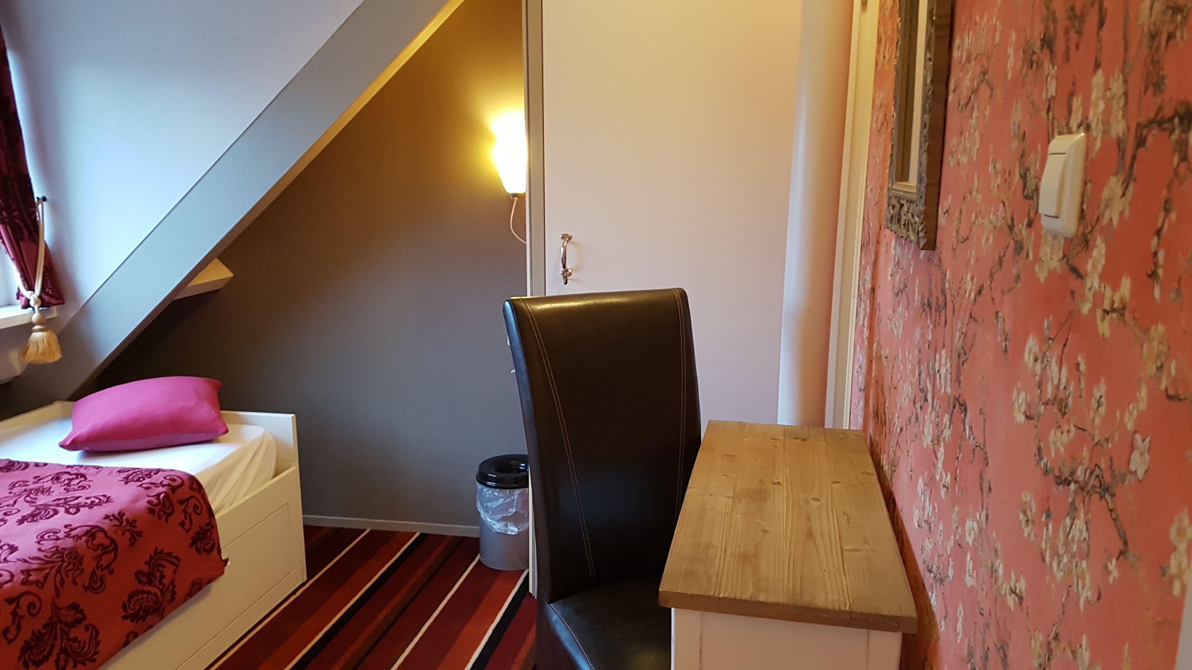 A small room with a bed, a chair, a wooden table, and a trash bin.