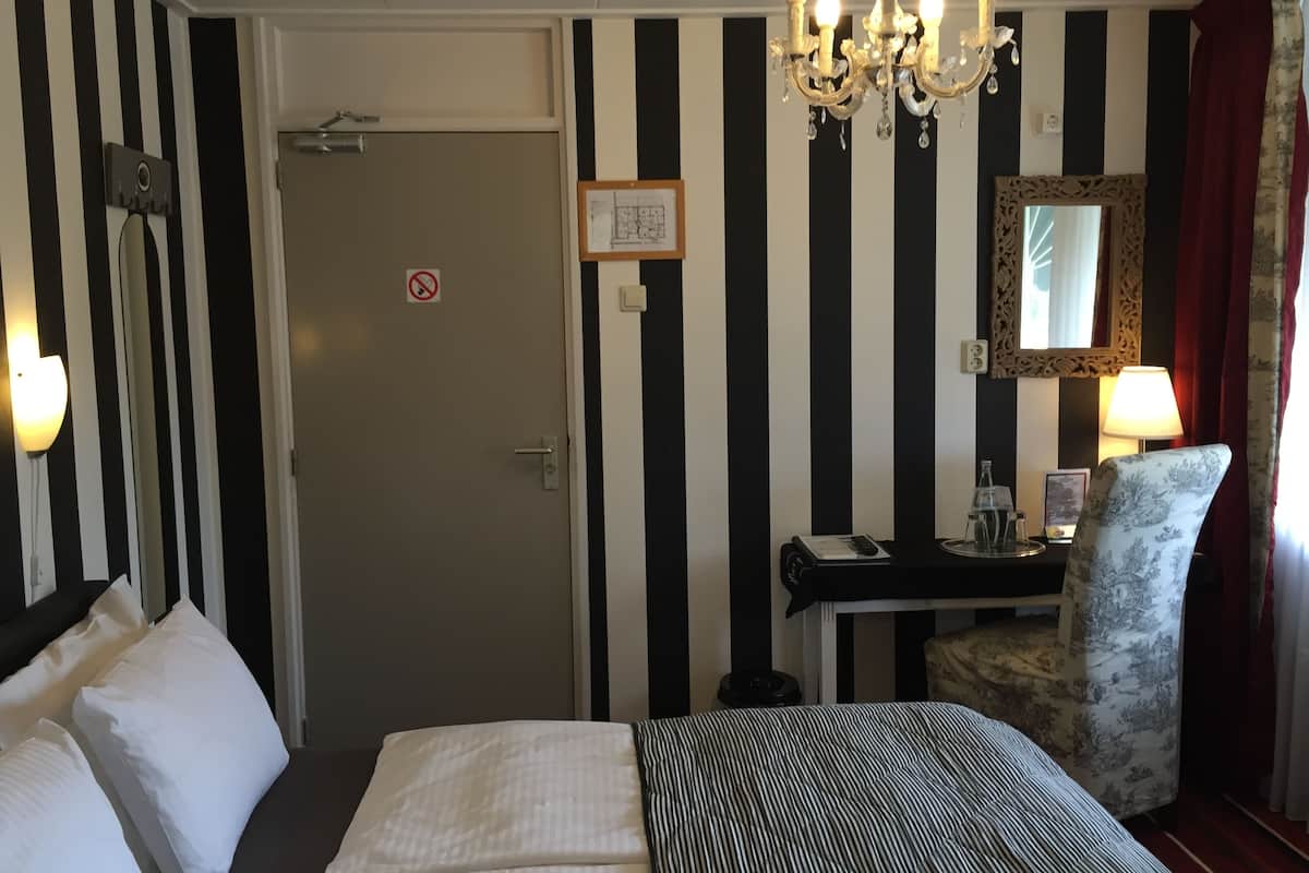 A hotel room with a bed, a desk, a chair, a chandelier, a mirror, and a door.