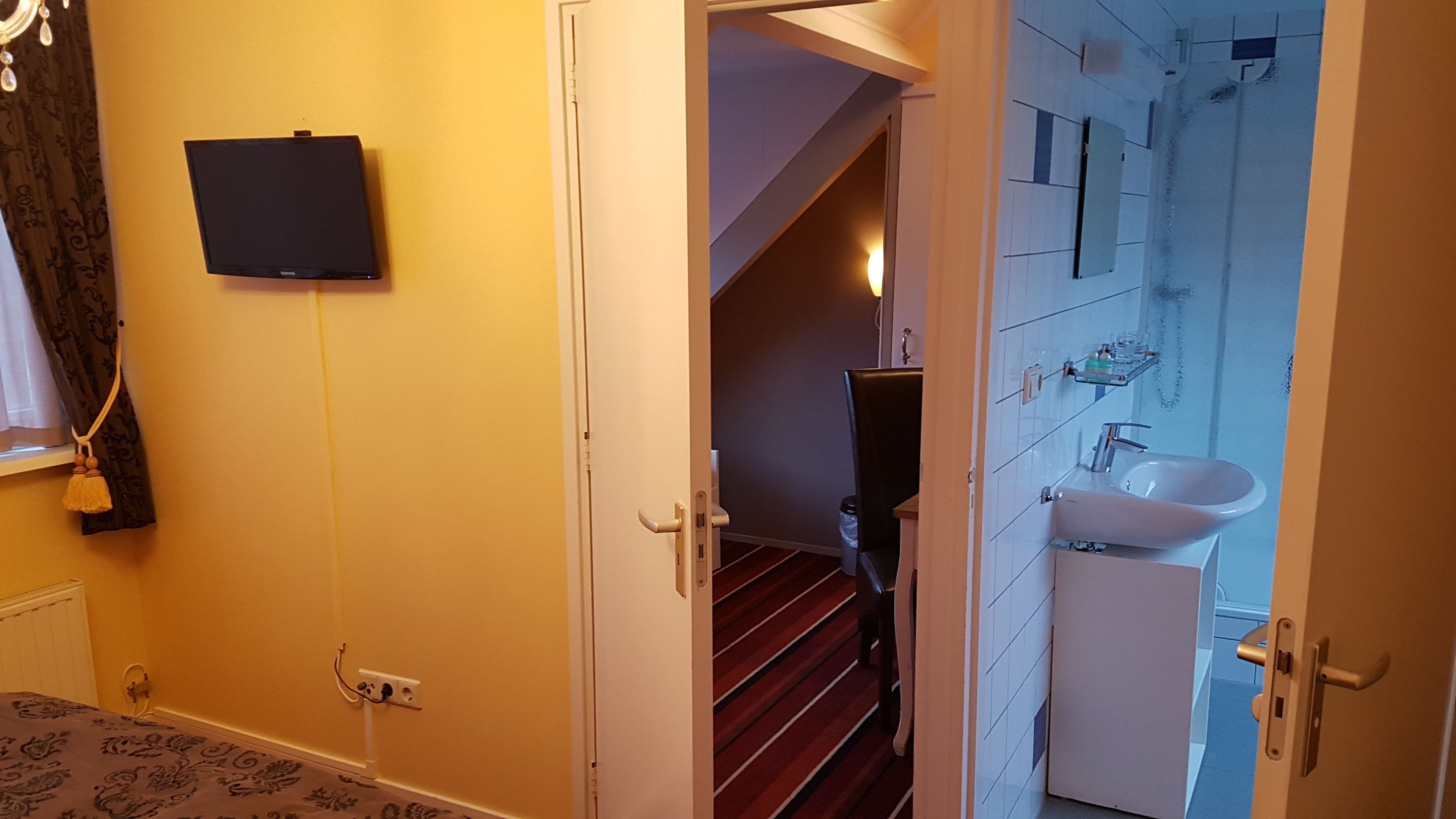 Une chambre comprenant un lit, un téléviseur à écran plat fixé au mur, une porte donnant sur une salle de bain avec un lavabo et un miroir, et un escalier.
