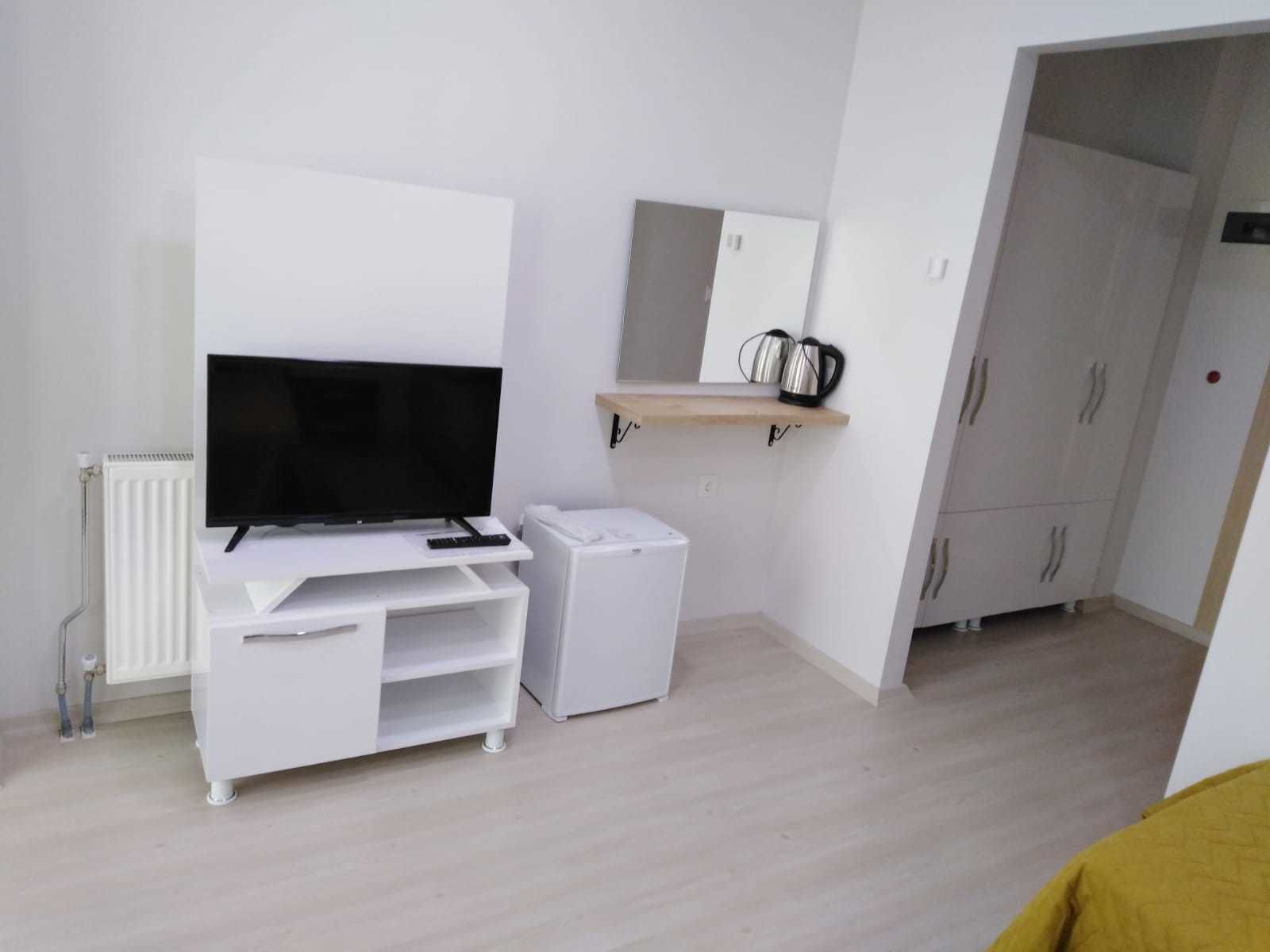 Ein modernes, minimalistisches Zimmer mit einem Flachbildfernseher auf einem weißen Standfuß, einem kleinen, weißen Kühlschrank, einem hölzernen Ablagefach mit einem Wasserkocher und weißen Schränken.