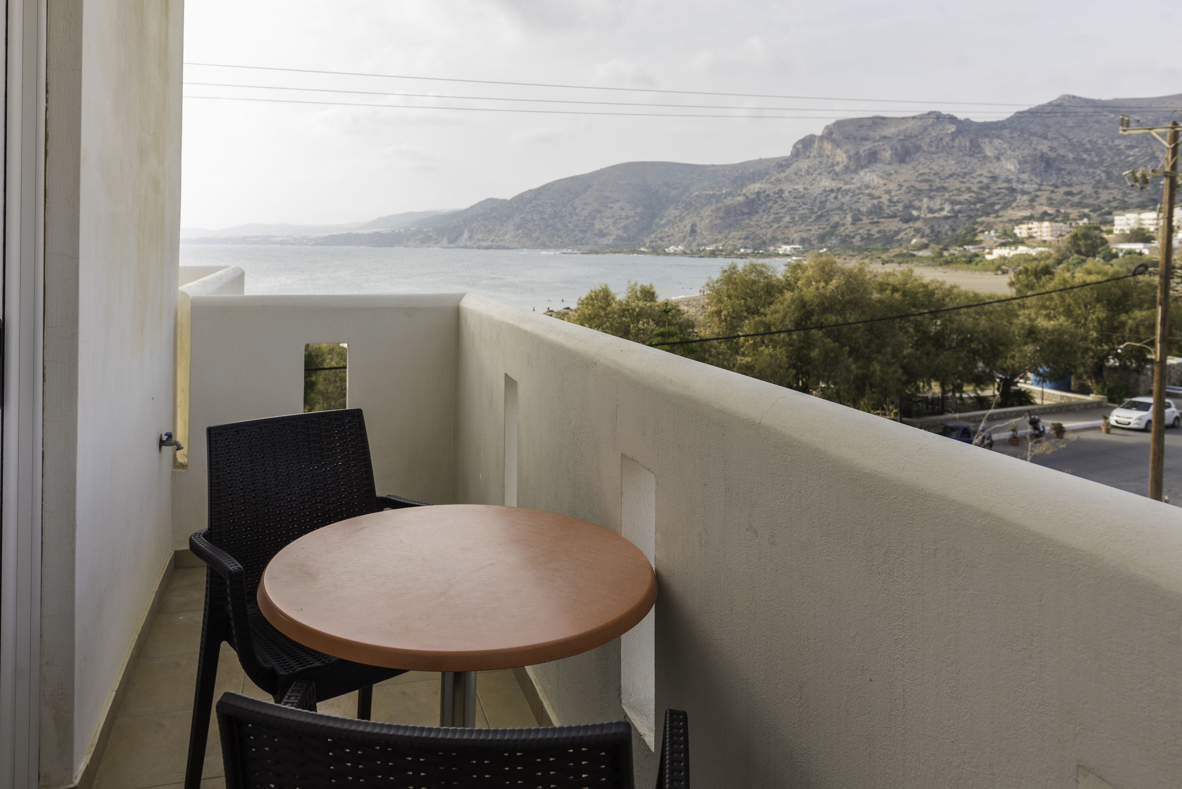 Un balcon avec une table ronde et deux chaises, donnant sur un paysage côtier avec des montagnes et une étendue d’eau.