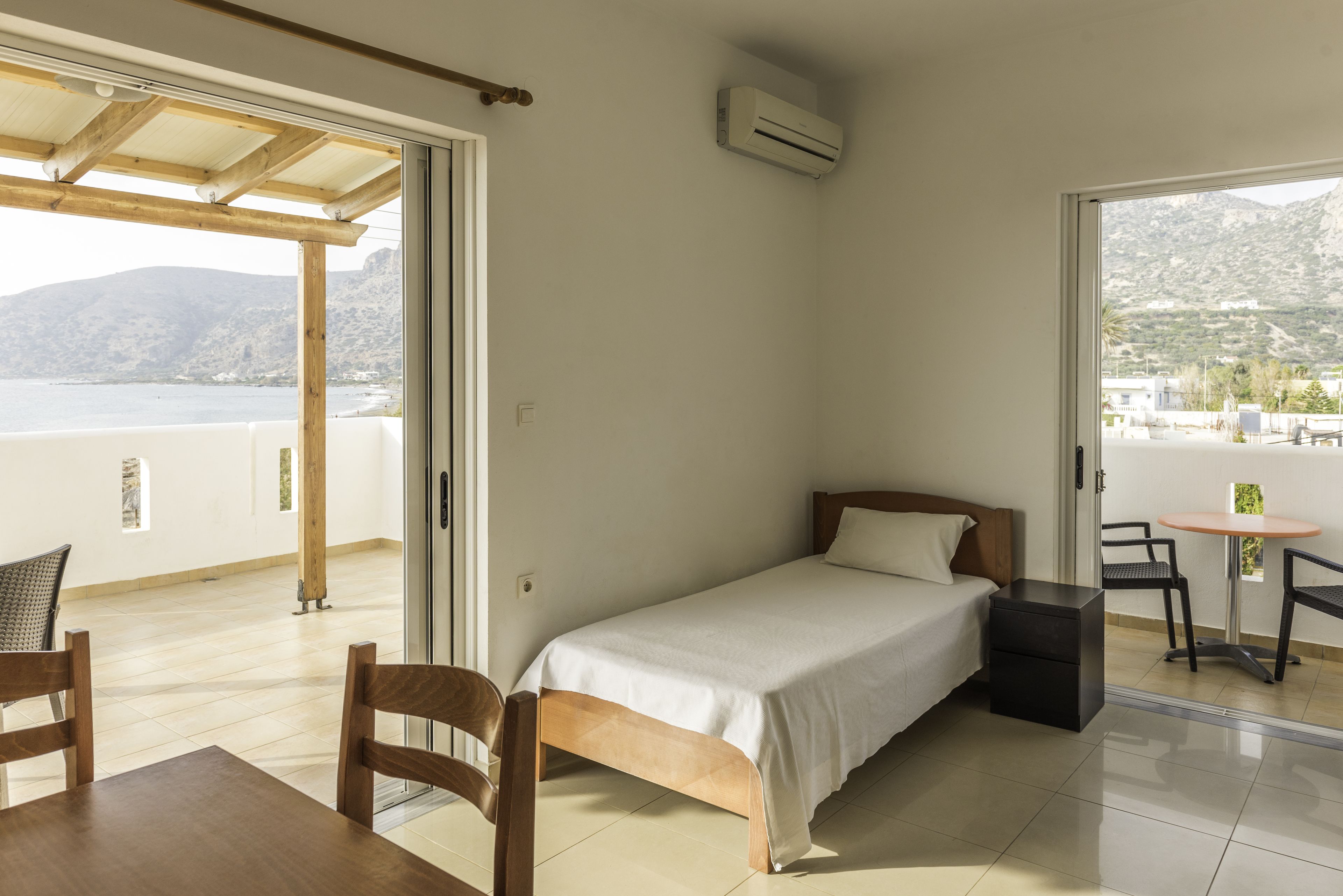 A bedroom with a bed, a table, and chairs, a sliding glass door leading to a balcony with a view of the sea and mountains.
