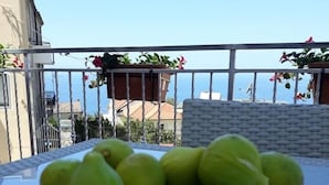On aperçoit clairement un balcon avec une table sur laquelle se trouve un saladier rempli de fruits verts. La vue donne sur une zone côtière avec des bâtiments et un ciel dégagé.