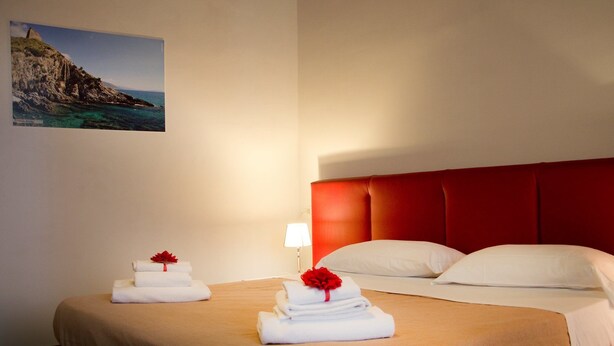 Une chambre d’hôtel avec un lit, deux serviettes ornées de fleurs rouges, une tête de lit rouge, des oreillers blancs et un tableau représentant un paysage côtier, accroché au mur.