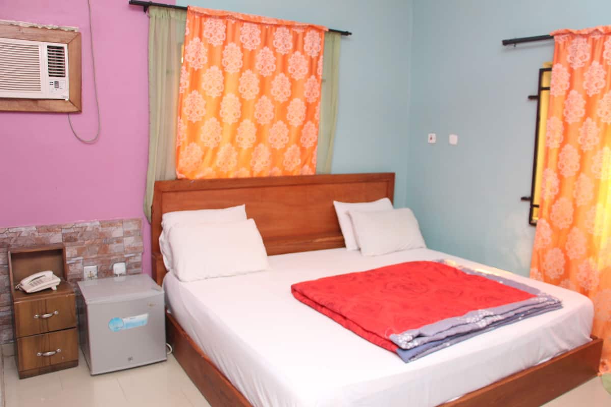 Un dormitorio con cama de madera, almohadas blancas, manta roja, aire acondicionado, mesita de noche y un armario pequeño.