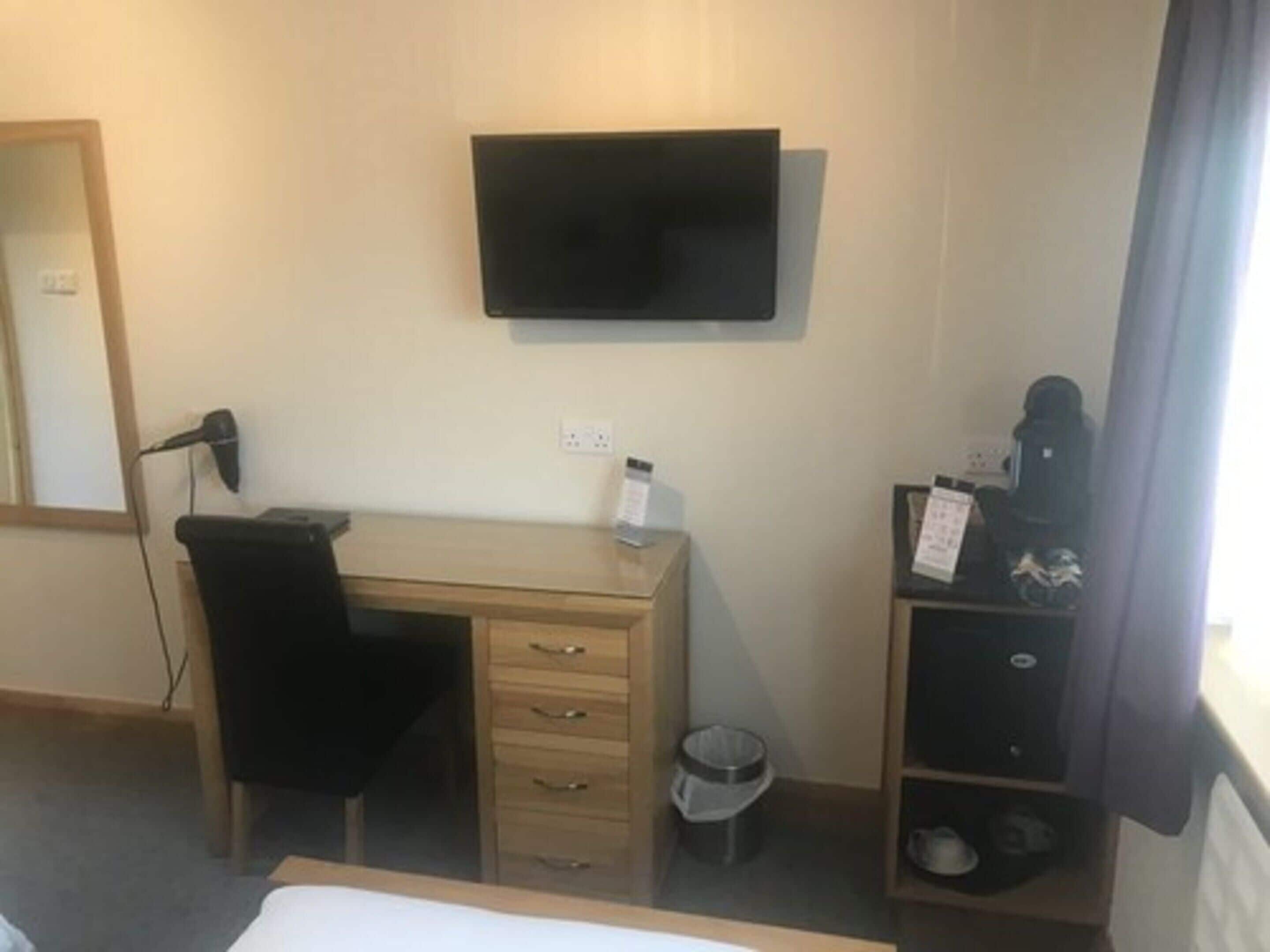 Ein Hotelzimmer mit einem Bett, einem Schreibtisch mit Stuhl, einem an der Wand befestigten Fernseher, einer kleinen Mülltonne und einem Schuhregal.