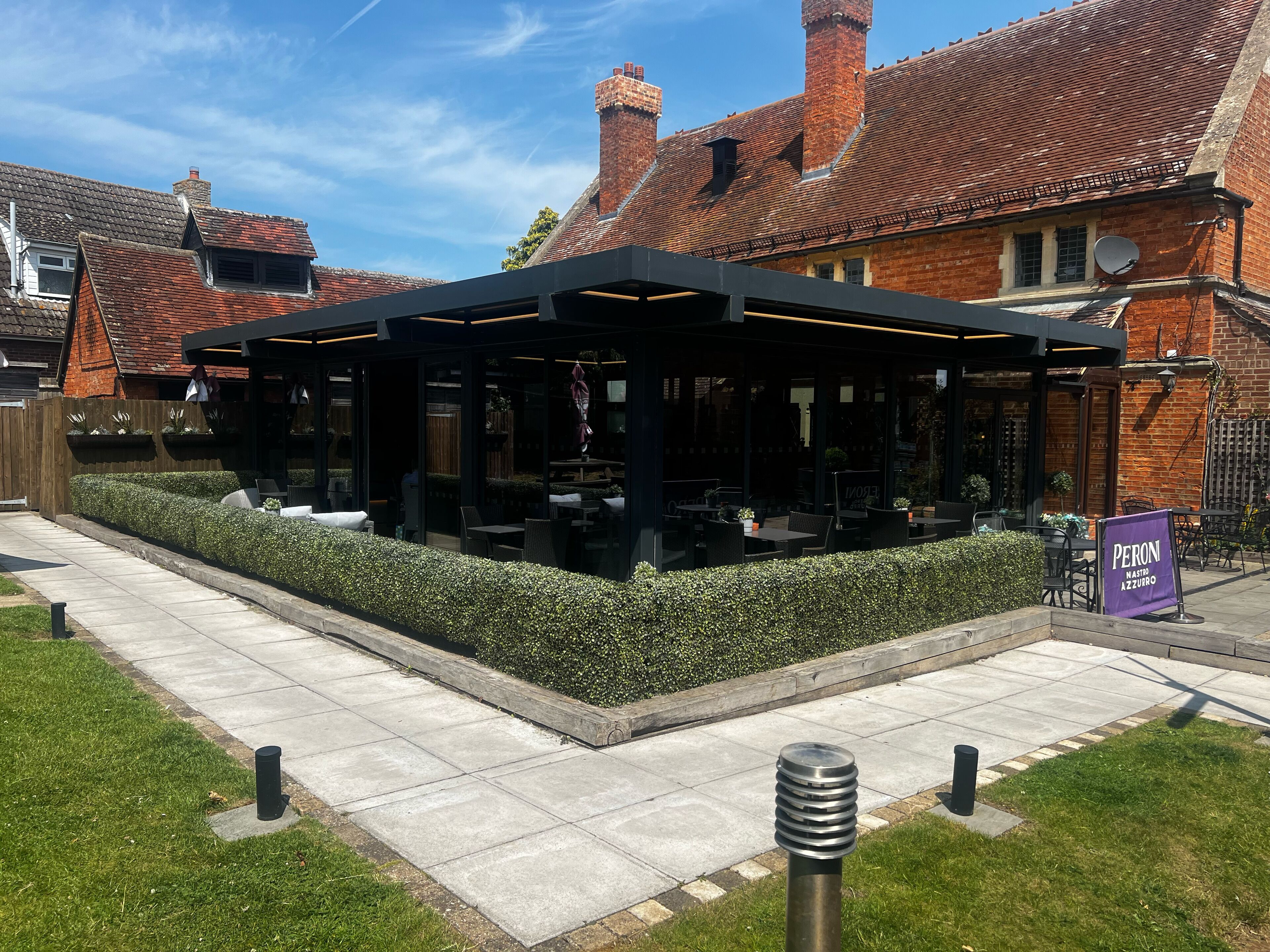 An outdoor seating area with tables and chairs, surrounded by a hedge and a brick building with a tiled roof.