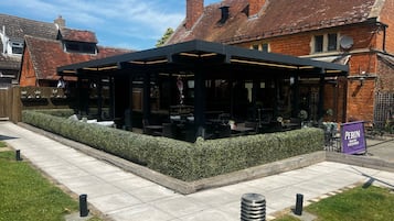 An outdoor seating area with tables and chairs, surrounded by a hedge and a brick building with a tiled roof.