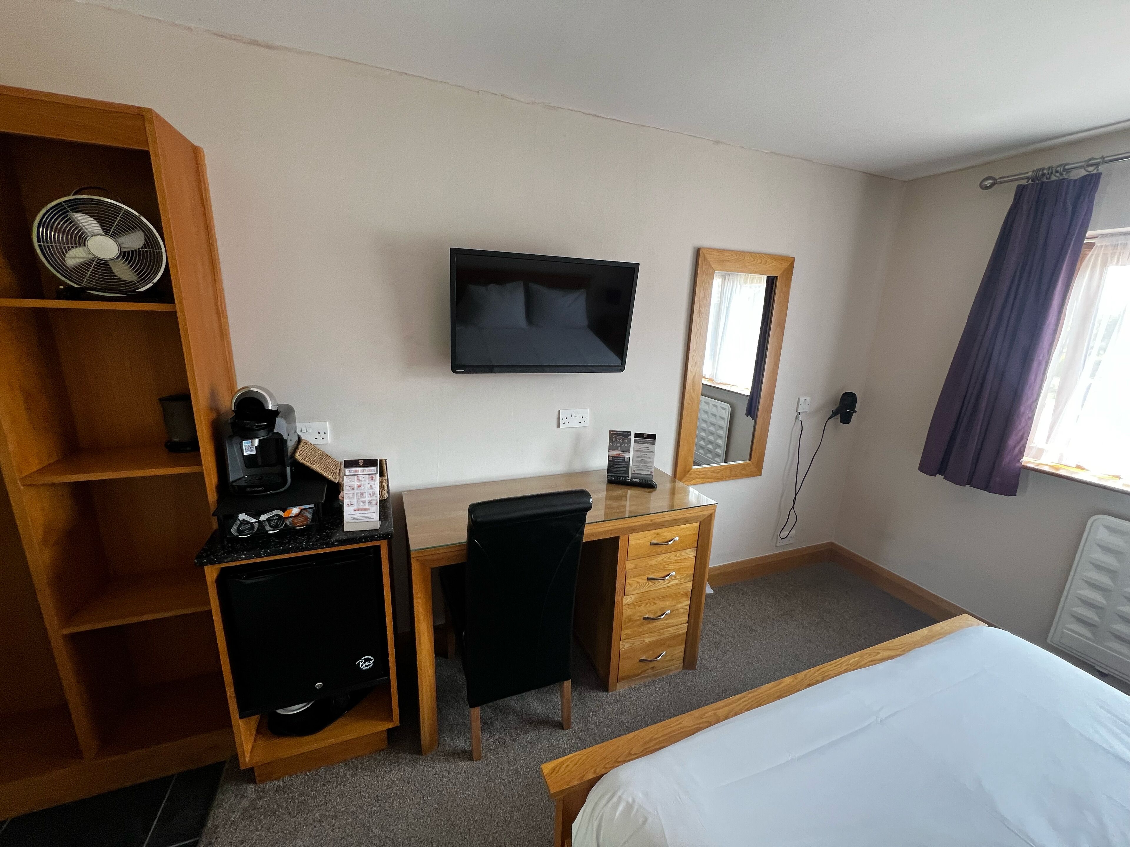 Een hotelkamer met een bed, een bureau met stoel, een televisie, een spiegel, een ventilator en een koffieapparaat.