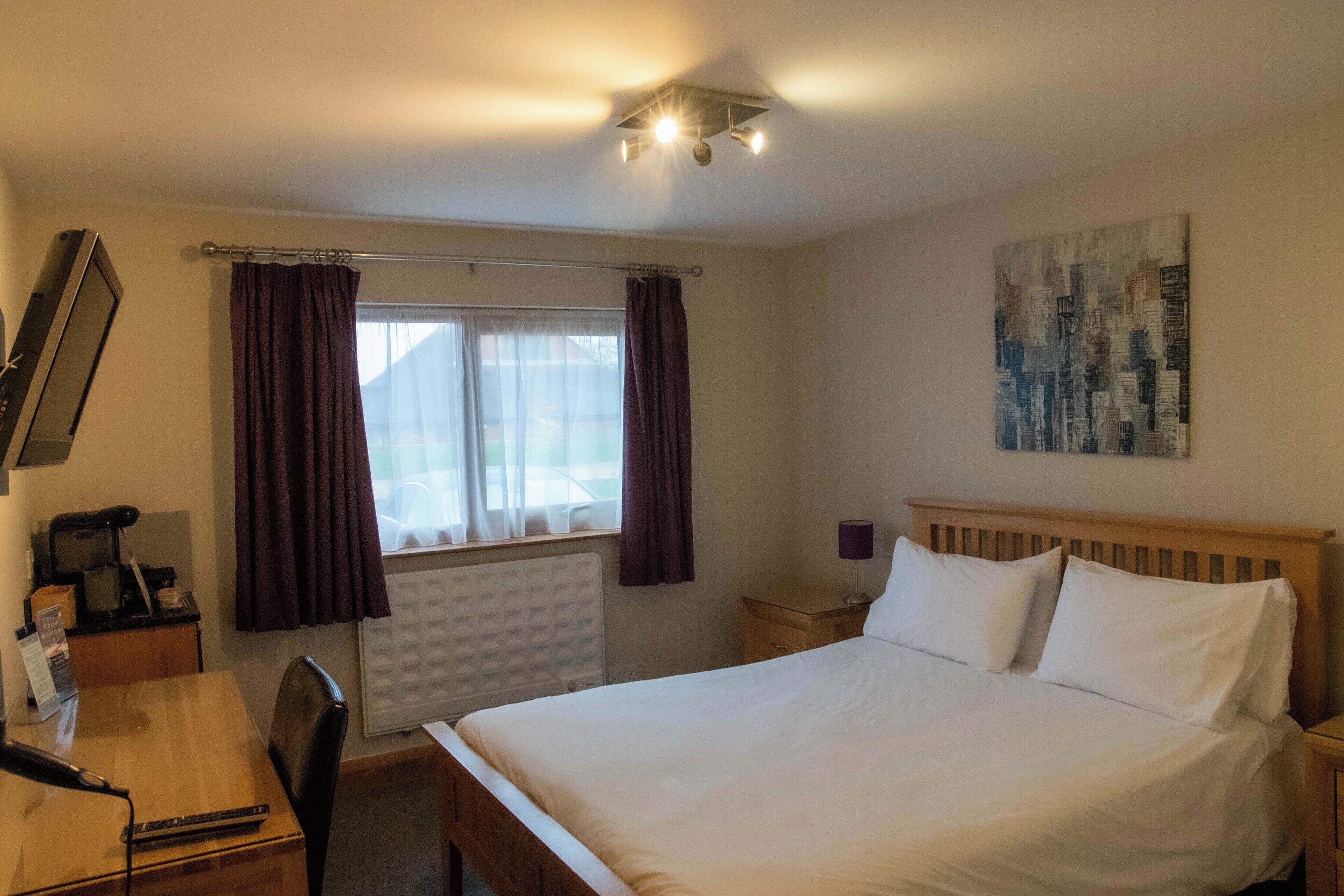 Habitación de hotel con cama, escritorio con silla, ventana con cortinas, radiador, mesita de noche con lámpara y una obra de arte colgada en la pared.
